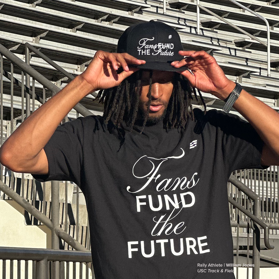 Person wearing a black t-shirt and cap with 'Fans Fund the Future' text, standing in bleachers.